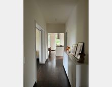 Ein hell beleuchteter Flur in einem Wohnhaus mit dunklem Holzboden und weißen Wänden. Links befinden sich zwei Türen, rechts steht ein weißes Sideboard mit Dekorationen, am Ende des Flurs sind eine offene Küche und ein Essbereich mit großen Fenstern sichtbar.