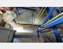 Blick in das Innere einer industriellen Anlage mit großen Rohrleitungen und silbernen Behältern. Die Konstruktion ist von gelben und blauen Streben sowie Metallgittern umgeben, mit sichtbaren Rohrleitungen und mechanischen Verbindungen.