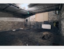 Ein leerer, alter Kellerraum mit Ziegelwänden, einer Betondecke und einem erdigen Boden. Im Raum befinden sich freiliegende Rohre an der Decke, eine gelbe Gasleitung an der Wand und einige beschädigte Bretter, die an der Wand angebracht sind.