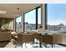 Moderner Sitzbereich mit cremefarbenen Sesseln und einem weißen Tisch, platziert vor großen bodentiefen Fenstern, die den Blick auf ein städtisches Dachpanorama ermöglichen. Helle Beleuchtung durch Tageslicht, mit einer dekorativen Blumenvase auf dem Tisch.