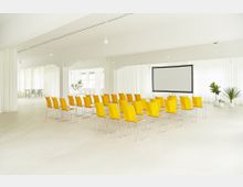 Konferenzraum mit gelben Stühlen in Reihen vor einer großen, weißen Projektionsleinwand. Der Raum ist hell, mit weißen Wänden, bodentiefen Fenstern und dekorativen Zimmerpflanzen.
