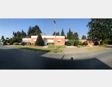 Straßenszene mit flachen, einstöckigen Wohnhäusern in einer ruhigen Wohngegend. Die Häuser sind umgeben von gepflegten Gärten, Zufahrten mit geparkten Autos und einer Laterne im Vordergrund; im Hintergrund sind mehrere hohe Bäume zu sehen.