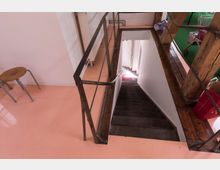Treppenabgang in einem Innenraum mit metallischem Geländer, einem hellem rosa Boden und Holzakzenten. Im Hintergrund sind Teile eines Raums mit grünem Teppich und abstellbaren Gegenständen zu sehen.