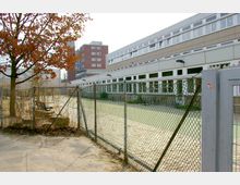 Schulgebäude mit langen, hellen Fassaden und vielen Fenstern, umgeben von einem Drahtzaun. Im Vordergrund ein Baum mit braunem Laub und ein leerer, betonierter Bereich. Im Hintergrund ein mehrstöckiges rotes Backsteingebäude.