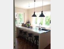 Moderne Küche mit einer weißen Kochinsel, an der vier schwarze Hocker stehen. Helle Fenster an zwei Seiten lassen viel Tageslicht herein, darüber hängen zwei schwarze Pendelleuchten; im Hintergrund sind Bäume sichtbar.