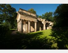 Historische Ruinen einer antiken Säulenhalle mit dekorativen Kapitellen, teilweise eingestürzt, umgeben von Bäumen und einer Wiese bei Tageslicht. Im Hintergrund dichter Baumbewuchs unter einem klaren, blauen Himmel.