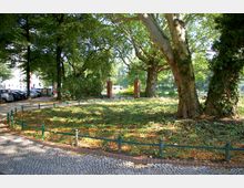 Stadtpark mit großer Rasenfläche, von niedrigen grünen Geländern umgeben, im Vordergrund ein gepflasterter Weg. Im Hintergrund sind hohe Bäume, zwei Backsteinsäulen mit Laternen und ein Zaun zu erkennen.
