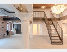 Ein offener Innenraum mit industriellem Charakter, geprägt von freiliegenden Backsteinwänden, einer weißen Säule, und einer Metalltreppe, die zu einer oberen Galerie führt. Eine große Kristallleuchte hängt von der Decke, und eine schwere Metalltür sowie Fenster sorgen für zusätzliche Akzente.