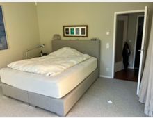 Schlafzimmer mit einem großen, grauen Polsterbett und weißer Bettdecke. Im Hintergrund ist eine geöffnete Tür zu sehen, daneben eine helle Wand mit Lichtschaltern, sowie an der linken Seite ein Kunstbild an der Wand und eine Nachttischlampe.