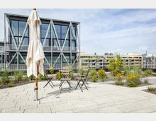 Dachterrasse mit Sitzgelegenheiten, Sonnenschirm und modernen Pflanzbeeten; im Hintergrund ein Gebäude mit Glasfassade und diagonalen Metallverstrebungen sowie weitere Stadtarchitektur.