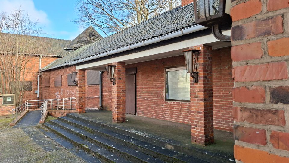 Backsteinbau mit überdachtem Eingangsbereich, getragen von gemauerten Pfeilern, davor breite Treppenstufen und eine Rampe mit Metallgeländer. An der Fassade sind Wandlampen, eine Tür und ein Fenster unter einem dunklen Ziegeldach zu sehen, im Hintergrund stehen kahle Bäume.
