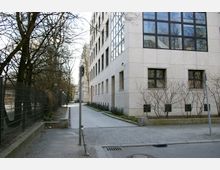 Ein moderner Gebäudekomplex mit heller Fassade und großen Fenstern, flankiert von einem Gehweg und einem Metallzaun auf der linken Seite. Karge Bäume und Sträucher säumen die Umgebung, die in einer urbanen Straße mit Pflastersteinen liegt.