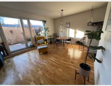 Wohnzimmer mit Holzboden, großen Glastüren, die zu einer Terrasse führen, und minimalistischer Einrichtung. Im Raum befinden sich ein gelber Sessel, ein runder Esstisch mit Stühlen, mehrere Pflanzen und hängende Lampen an der Decke.