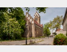 Backsteinkirche mit roten Dächern und facettenreichen Gotik-Elementen, eingebettet in eine grüne Umgebung mit Kastanienbäumen. Die Pflasterstraße im Vordergrund führt an der Kirche vorbei, mit einigen Laternen und Gebäuden im Hintergrund.