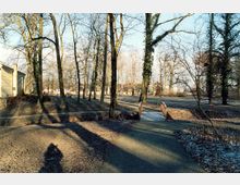 Städtischer Park mit laubfreien Bäumen im Winter, einem kleinen Holzsteg über einen Graben und Wegen, die sich durch die Anlage schlängeln. Rechts im Bild sind Wiesen und Sträucher erkennbar, links ein Gebäude am Rand des Parks.