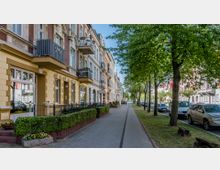 Straßenansicht mit historischen Wohnhäusern im Gründerzeitstil, links gelb und rot verklinkerte Fassaden mit Balkonen, rechts eine von Bäumen gesäumte Allee mit parkenden Autos und einem Gehweg.