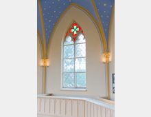 Kirchenfenster im neugotischen Stil mit einer farbigen Rosette aus rotem und grünem Glas im oberen Bereich und darunter verglasten Sprossenelementen. Der Innenraum ist durch eine blaue, sternverzierte Decke und helle Wände mit dekorativen Säulen geprägt.