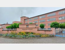 Rosa Schulgebäude der Friedrich-Ebert-Grundschule mit mehreren großen Fenstern und einer Uhr am Turm. Im Vordergrund sind ein bepflanztes Blumenbeet, eine niedrige Mauer und blaue Zäune zu sehen.