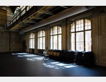 Großer, leerer Industriehallenraum mit rauen Betonwänden und hoher Decke mit sichtbaren Stahlträgern und Lüftungskanal. Entlang einer Wand stehen hohe, bogenförmige Sprossenfenster, durch die Licht auf den Boden fällt; darunter sind gestapelte Klappstühle angeordnet.