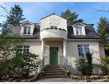 Zweistöckiges Einfamilienhaus mit hellem Putz, klassischem Satteldach und zwei Gaubenfenstern. Eine Eingangstür mit grünem Anstrich ist über eine Treppe zugänglich, eingerahmt von weißen Säulen, darüber ein kleiner Balkon mit Pflanzen.