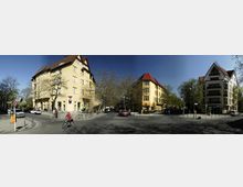 Straßenkreuzung in einem Wohngebiet mit mehrstöckigen Gebäuden, darunter gelbe Häuser mit roten Dächern im traditionellen Stil und ein modernes Gebäude mit Balkonen. Die Szene zeigt parkende Autos, Radfahrer, Fußgänger und einige Bäume entlang der Straße bei klarem Himmel.