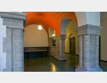 Historischer Innenraum mit gewölbten Decken und massiven Steinsäulen, die mit geschnitzten Mustern verziert sind. Eine Sitzbank steht an einer Wand, und die Deckenbögen sind in einem auffälligen Orangeton gefärbt, während Hängelampen den Raum beleuchten.