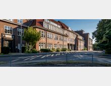 Backstein-Gebäude im Stil der klassischen Architektur mit großen Fenstern, rotem Satteldach und einer auffälligen Uhr über einem Eingang. Das Gebäude liegt an einer ruhigen Straße, von Bäumen und Gehwegen gesäumt.