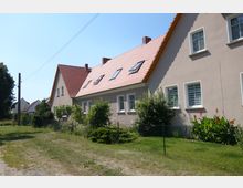 Zwei Reihenhäuser mit modernen, beigefarbenen Fassaden und roten Satteldächern, umgeben von gepflegten Gärten mit Sträuchern und Blumen. Ein grüner Zaun trennt die Häuser vom Vorgarten, im Hintergrund sind Bäume und weitere Gebäude sichtbar.