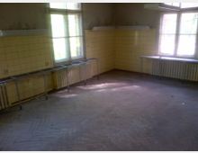 Ein leerer Raum mit gelb gefliesten Wänden zur Hälfte der Höhe, großen Fenstern, Heizkörpern und Metallgestellen entlang der Wände. Der Boden besteht aus Holz in Fischgrätmuster, und der Raum ist spärlich beleuchtet.