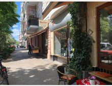 Straßenszene mit einer Reihe von Altbauwohnungen mit Balkonen auf der linken Seite und einem Antiquariat mit einem Schild "Bücher" auf der rechten Seite. Vor dem Geschäft stehen Bücherkisten, daneben rankt Efeu an der Fassade, und ein kleiner Tisch mit Stühlen steht am Gehweg.
