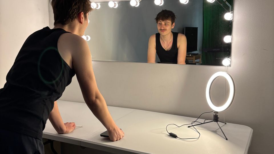 Small dressing-room style vanity area with a long wall mirror framed by bright round bulbs above a white folding table. A lit ring light on a tripod sits on the tabletop, with a wooden bench below and plain white walls surrounding the setup.
