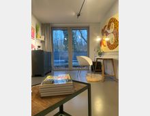 Kleines, modernes Büro mit großen Fensterfronten, die den Blick auf Bäume und einen Zaun freigeben. Im Raum steht ein schlicht gestalteter Schreibtisch mit einem weißen Stuhl und einem farbenfrohen Wandbild; im Vordergrund liegt ein Stapel Zeitschriften auf einem niedrigen Tisch.