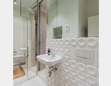 Modernes Badezimmer mit weißen Wandfliesen in geometrischem Kreisdesign, einem kleinen Waschbecken mit Spiegel darüber und Glasdetails. Im Hintergrund sind ein schmaler Wandspiegel, eine Duschkabine und ein heller Fliesenboden sichtbar.