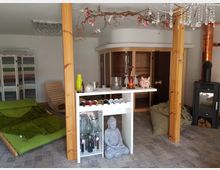 Ein Wellnessraum mit einer Sauna in Holzoptik, einem Kamin mit Kupferschornstein und gemütlichen Sitzgelegenheiten, darunter eine grüne Liegefläche. Im Vordergrund steht ein weißes Regal mit einer Getränkestation, Weinflaschen und dekorativen Elementen, darunter eine Buddha-Statue.