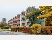 Wohnhaus in einer Wohnsiedlung, bestehend aus mehreren Etagen mit pastellfarbenen Balkonen in Rosa und Grün. Vor dem Gebäude befinden sich Gartenflächen mit Büschen, Bäumen und einem Gehweg, umgeben von herbstlicher Vegetation.