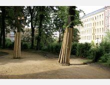 Ein kleiner Park mit hohen Bäumen, von denen einige mit Holzplanken abgestützt sind. Im Hintergrund sind mehrstöckige Wohn- und Geschäftshäuser mit hellen Fassaden zu sehen.