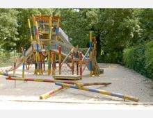 Ein Spielplatz mit Holzspielgeräten, darunter ein großer, bunter Kletterturm mit Rutschen, Netzen und Balken. Der Boden ist mit Sand bedeckt, umgeben von Bäumen und einer Hecke.