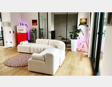 Modernes Wohnzimmer mit hellem Holzboden, einer cremefarbenen, wellenförmigen Couch und einem bunten, runden Teppich. Im Hintergrund ein roter Retro-Kühlschrank, ein großer Pflanzenkübel, eine leuchtende Deko-Buchstabe „A“ sowie Glaswände, die einen Ausblick in einen angrenzenden Raum geben.