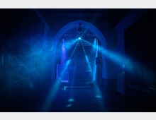 Ein dunkler Raum mit einem beleuchteten Durchgangsbogen, blauen Scheinwerferstrahlen und Nebeleffekten, typisch für einen Nachtclub oder Veranstaltungsraum. Im Hintergrund sind Lautsprecher und Bühnenbeleuchtung erkennbar.