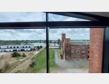 Blick durch ein großes Fenster auf eine Landschaft mit einem weißen, modernen Gebäudekomplex links, einem Backsteingebäude im historischen Stil rechts und einem Fluss in der Bildmitte, umgeben von einer grünen Wiesenlandschaft unter bewölktem Himmel.