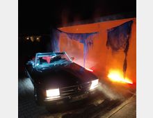 Ein schwarzes Cabriolet mit eingeschalteten Scheinwerfern steht auf einem gepflasterten Platz. Im Hintergrund ist eine orange beleuchtete Wand mit blau drapierten Stoffen und Nebeleffekten zu sehen.