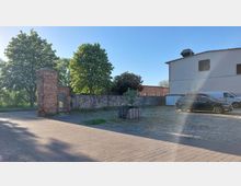 Ein gepflasterter Hof mit einer niedrigen Steinmauer, einem Tor aus Ziegeln und Holz, sowie einem kleinen Baum in einem quadratischen Holzbehälter. Im Hintergrund stehen blühende Bäume und ein Gebäude mit hellen Wänden, daneben geparkte Autos.