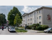 Mehrfamilienhaus aus der Nachkriegszeit mit schlichtem Putz, mehreren rechteckigen Fenstern und einer vertikalen Fensterfront im Eingangsbereich. Links führt eine Straße mit parkenden Autos und Bäumen entlang, im Vordergrund ein Gehweg mit Grünflächen und einer Person mit Gepäck.