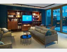 Wohnzimmer mit modernem Design: Ein Bücherregal aus Holz mit integriertem Gaskamin und einem Wandbildschirm dominiert den Raum. Graue Sofas, Couchtische mit Glasoberflächen und ein Sitzbereich mit Esstisch stehen vor großen Glaswänden, die auf eine Terrasse mit Pflanzen blicken.