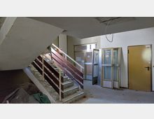 Treppenhaus eines älteren Gebäudes mit Betonstufen, Holzgeländer und gelben Innentüren. Der Raum zeigt Anzeichen von Renovierungsbedarf, mit abmontierten Türrahmen und Werkzeugen am Boden.