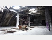 Verlassener Innenraum eines Gebäudes mit schrägen Betonwänden und mehreren runden Oberlichtern in der Decke, teilweise beschädigt. Im Vordergrund steht ein großer, rostiger Metallbehälter mit vertrockneten Ästen darin, umgeben von Schutt und Graffiti an den Wänden.