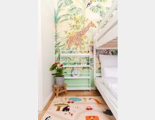 Kinderzimmer mit Etagenbett, bunter Dschungeltapete mit Tieren wie Giraffen und Zebras an der Wand, und einem hellen Kinderteppich mit Tier- und Fahrzeugmotiven auf Laminatboden. Links steht ein kleiner, hölzerner Hocker mit einer Topfpflanze darauf vor einem weißen Regal.