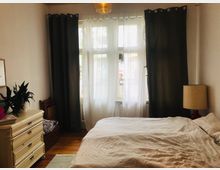 Ein Schlafzimmer mit einem großen Bett, weißer Tagesdecke und Holzboden. Im Hintergrund befindet sich ein Fenster mit weißen und dunklen Vorhängen, daneben stehen eine Stehlampe, eine cremefarbene Kommode mit einer Pflanze darauf und ein Holzstuhl.