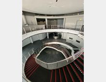 Ein modernes, zweistöckiges Innentreppenhaus mit spiralförmigem Design und Metallgeländern. Der Boden besteht aus dunklem Material, die Treppenstufen sind schwarz mit roten Streifen, und die Wände sowie die Decke sind weiß gehalten.