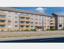 Vierstöckige Wohngebäude im schlichten Plattenbaustil mit beige-gelben Fassaden, zahlreichen Fenstern und Balkonen. Im Vordergrund befinden sich eine Straße, ein Gehweg und eine Bushaltestelle mit Werbeplakat.